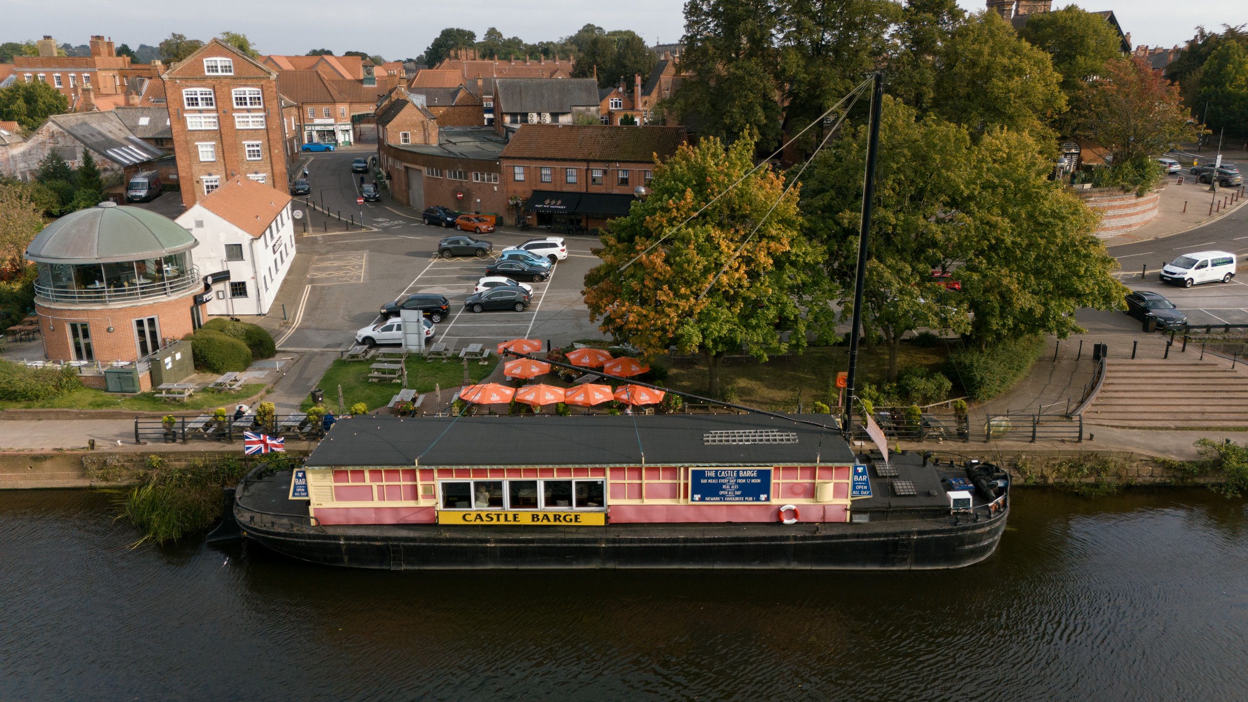 Aerial photograph of a large barge moored beside a riverside garden and brick buildings